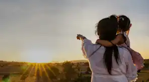 Frau mit Kind im Arm zeigt und blickt in Richtung aufgehende Sonne
