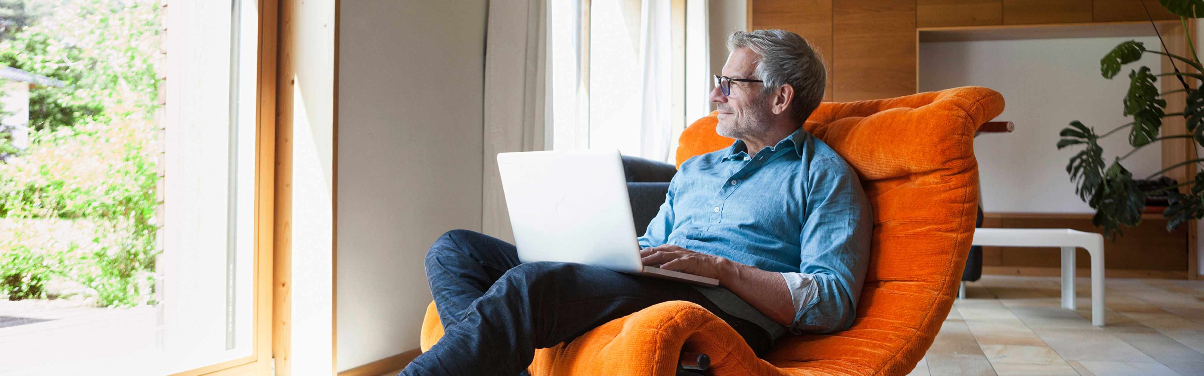 Älterer, zufriedener Mann in seinem Zuhause auf einem orangenem Sessel mit Laptop.