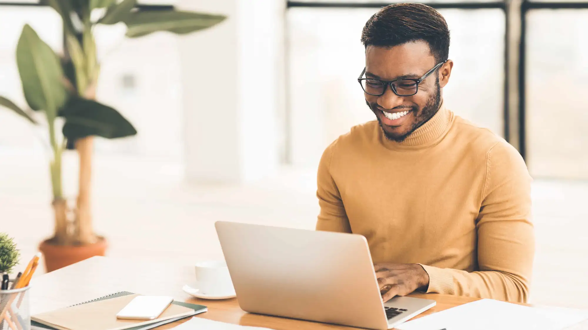 Junger Mann mit Brille lacht Laptop am Schreibtisch an mit Pflanze im Hintergrund