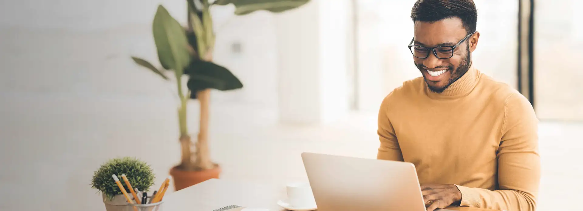 Junger Mann mit Brille lacht Laptop am Schreibtisch an mit Pflanze im Hintergrund