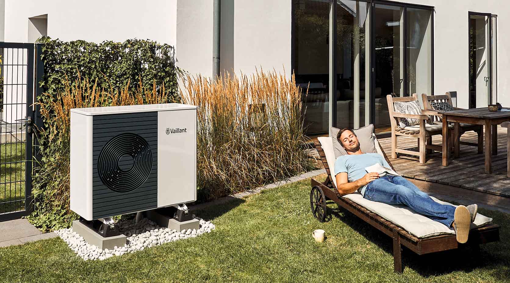 Mann liegt gemütlich auf einer Liege vor seinem Haus in der Nähe einer Wärmepumpe von Vaillant