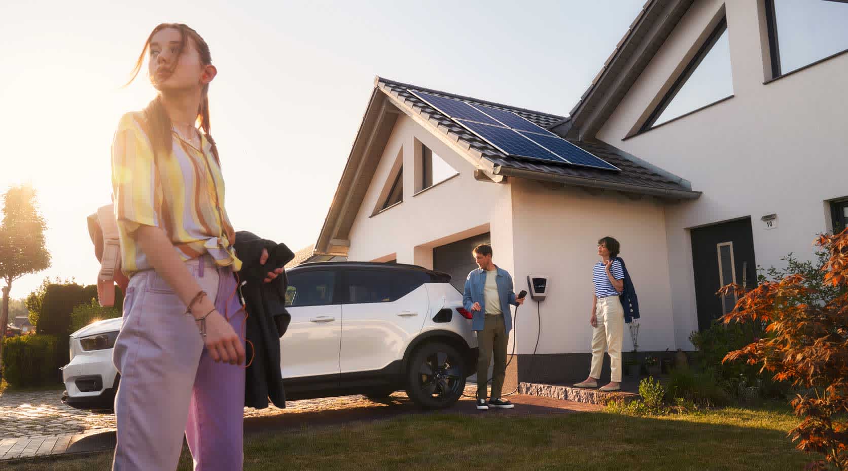 Mädchen mit ihren Eltern vor dem Haus und einem E-Auto, das an der Wallbox Strom vom eigenen Dach lädt