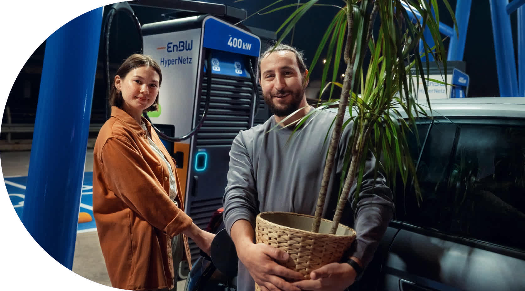 Ein Pärchen lädt an einer EnBW Ladesäule. Der Mann trägt eine Pflanze.