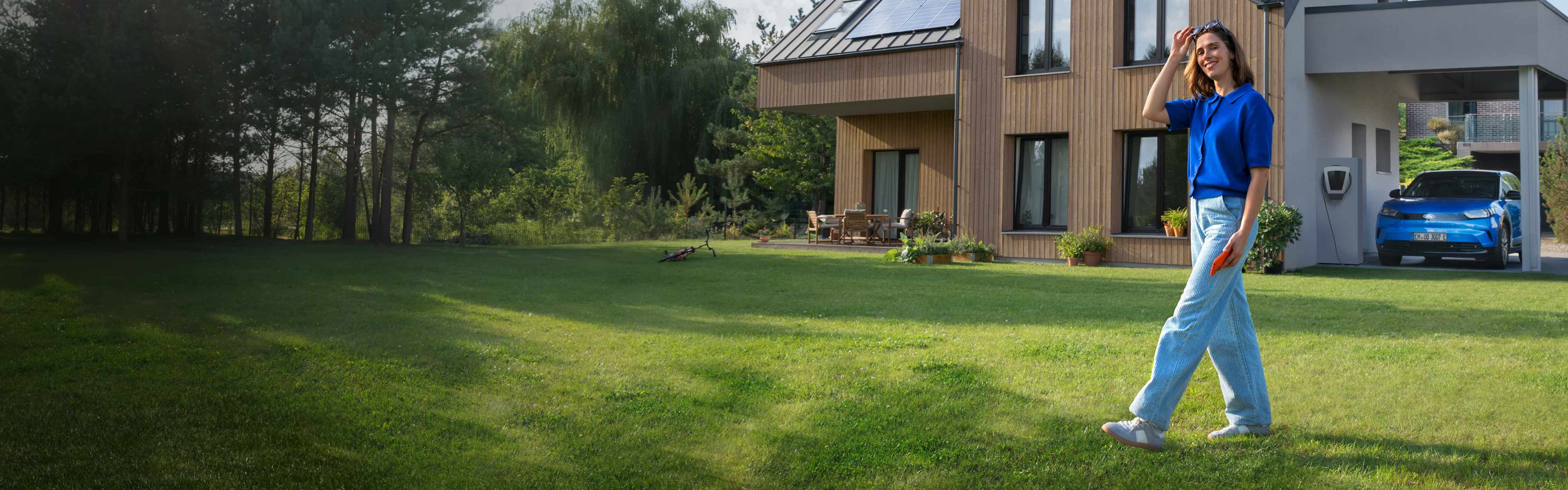 Eine Frau läuft durch den Garten ihres Hauses, im Hintergrund lädt das E-Auto mit Solarstrom vom Hausdach.