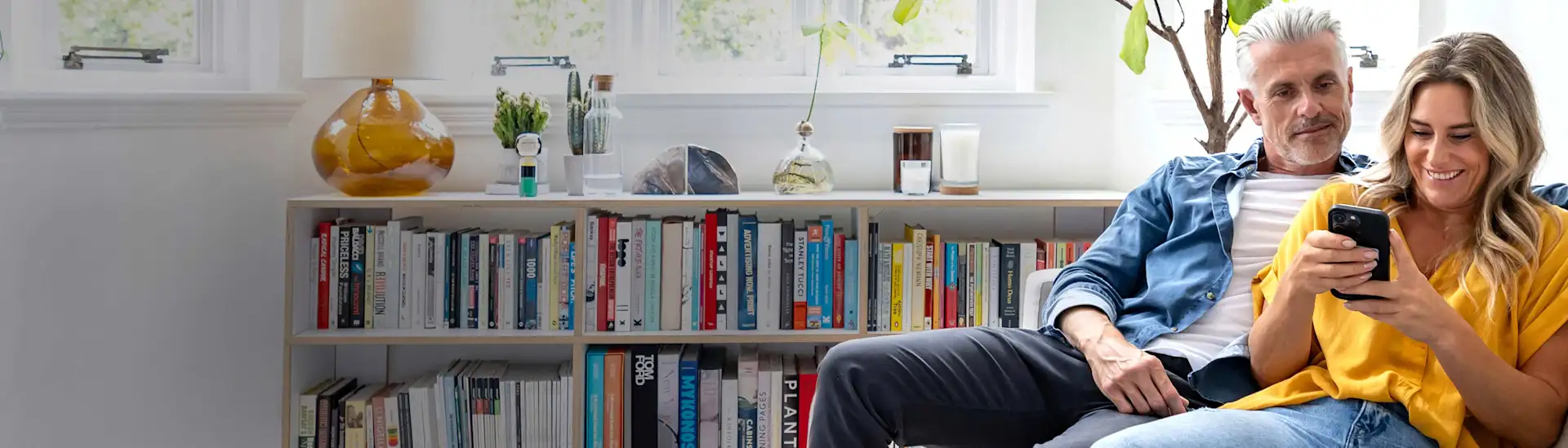 Paar im mittleren Alter sitzt auf der Couch und schaut auf ein Smartphone.