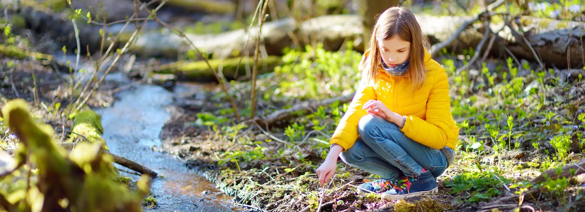 Mädchen in gelber Jacke spielt im Wald am Fluss Stock