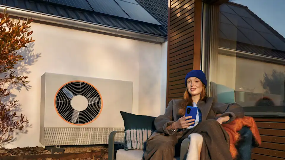 KI-generiert: Eine Frau sitzt im Garten auf einer Bank mit vielen Decken und Kissen. Im Hintergrund sind eine Holzhausfassade und eine Wärmepumpe zu sehen.