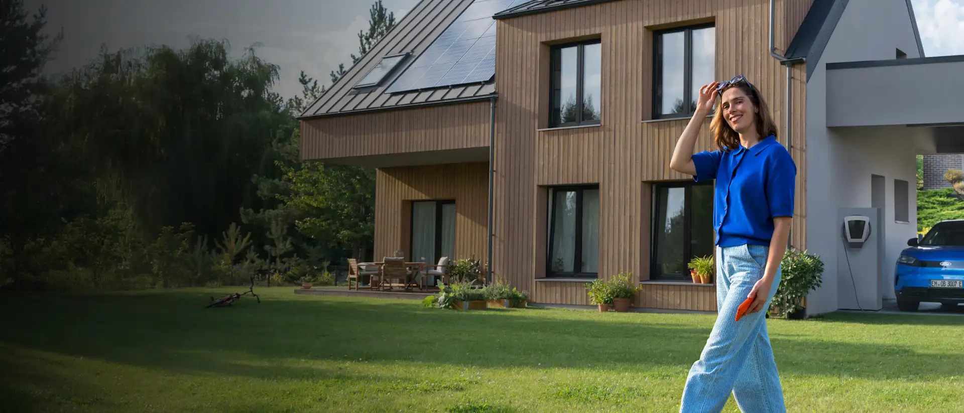 Eine Frau läuft durch den Garten ihres Hauses, im Hintergrund lädt das E-Auto mit Solarstrom vom Hausdach.