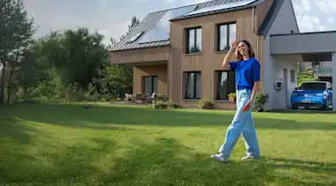 Eine Frau läuft durch den Garten ihres Hauses, im Hintergrund lädt das E-Auto mit Solarstrom vom Hausdach.