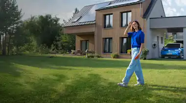 Eine Frau läuft durch den Garten ihres Hauses, im Hintergrund lädt das E-Auto mit Solarstrom vom Hausdach.
