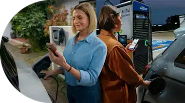 Zwei Frauen laden ihr Auto zuhause und unterwegs und prüfen den Ladevorgang in der EnBW mobility+ App auf ihrem Smartphone