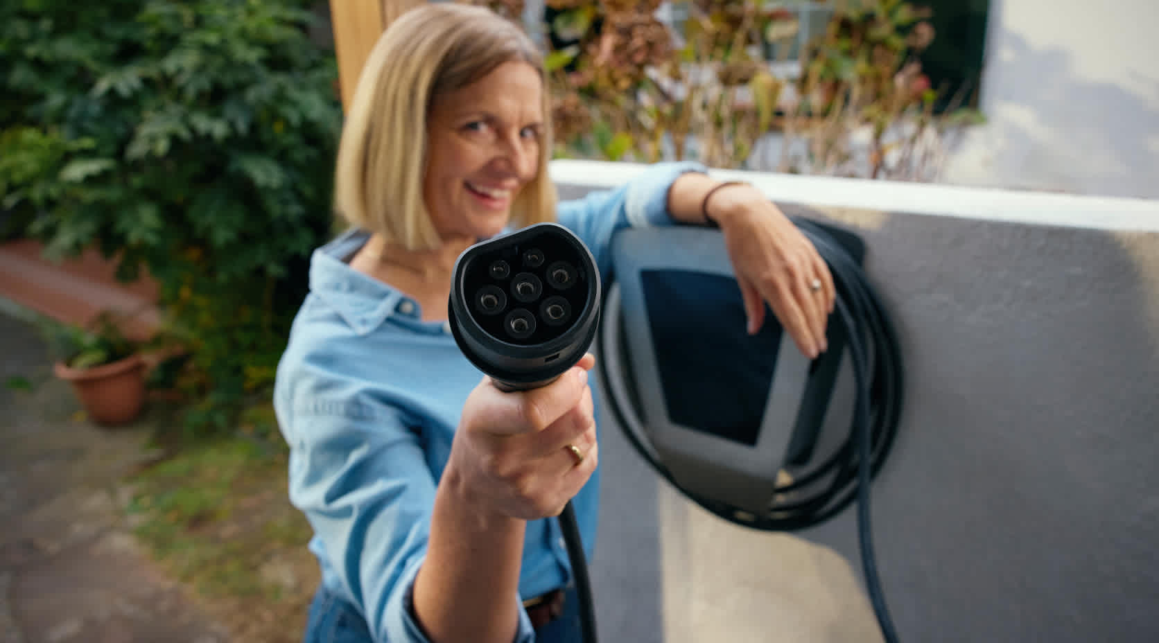 Ein Frau steht an einer Wallbox und hält den Stecker in die Kamera.