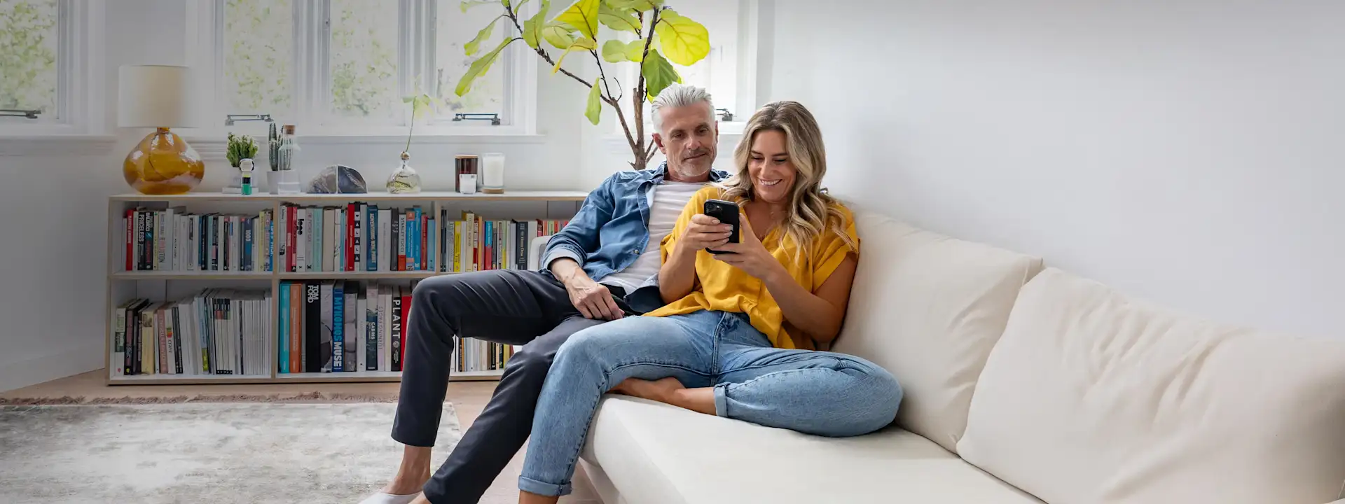 Paar im mittleren Alter sitzt auf der Couch und schaut auf ein Smartphone.
