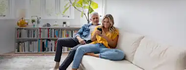 Paar im mittleren Alter sitzt auf der Couch und schaut auf ein Smartphone.