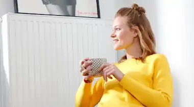Junge Frau mit gelbem Pullover und Tasse sitzt vor Heizung Fenster