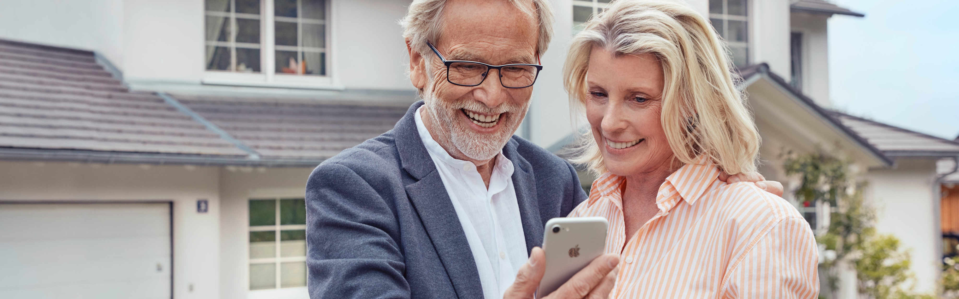 Best-Ager-Paar mit Smartphone vor eigenem Haus