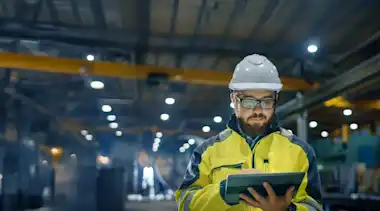 Mann mit Tablet steht in einer Lagerhalle, er trägt Schutzhelm und Schutzkleidung