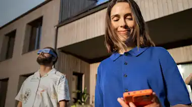 Eine Frau steht vor ihrem Haus, wirft einen Blick auf EnBW Mavi und lächelt. Hinter ihr steht ein Mann mit einer Sonnenfinsternis-Brille.