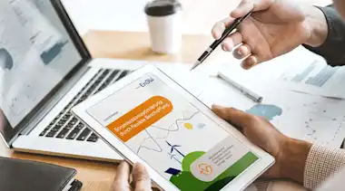 Hände halten Tablet mit Whitepaper "Stromkostenoptimierung durch flexible Beschaffung" neben Laptop