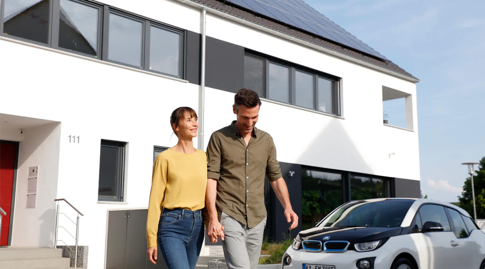 Paar vor einem Eigentumshaus mit einer Solaranlage und einem E-Auto davor