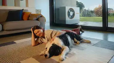 Junge Frau liegt mit ihrem Hund auf einem Teppich im Wohnzimmer ihres Hauses. Im Hintergrund auf der Terrasse ist eine Wärmepumpe  zu sehen.