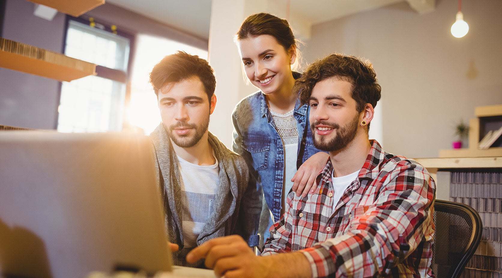 Zwei junge Männer und eine junge Frau schauen auf einen Monitor und nehmen am E-Learning teil.