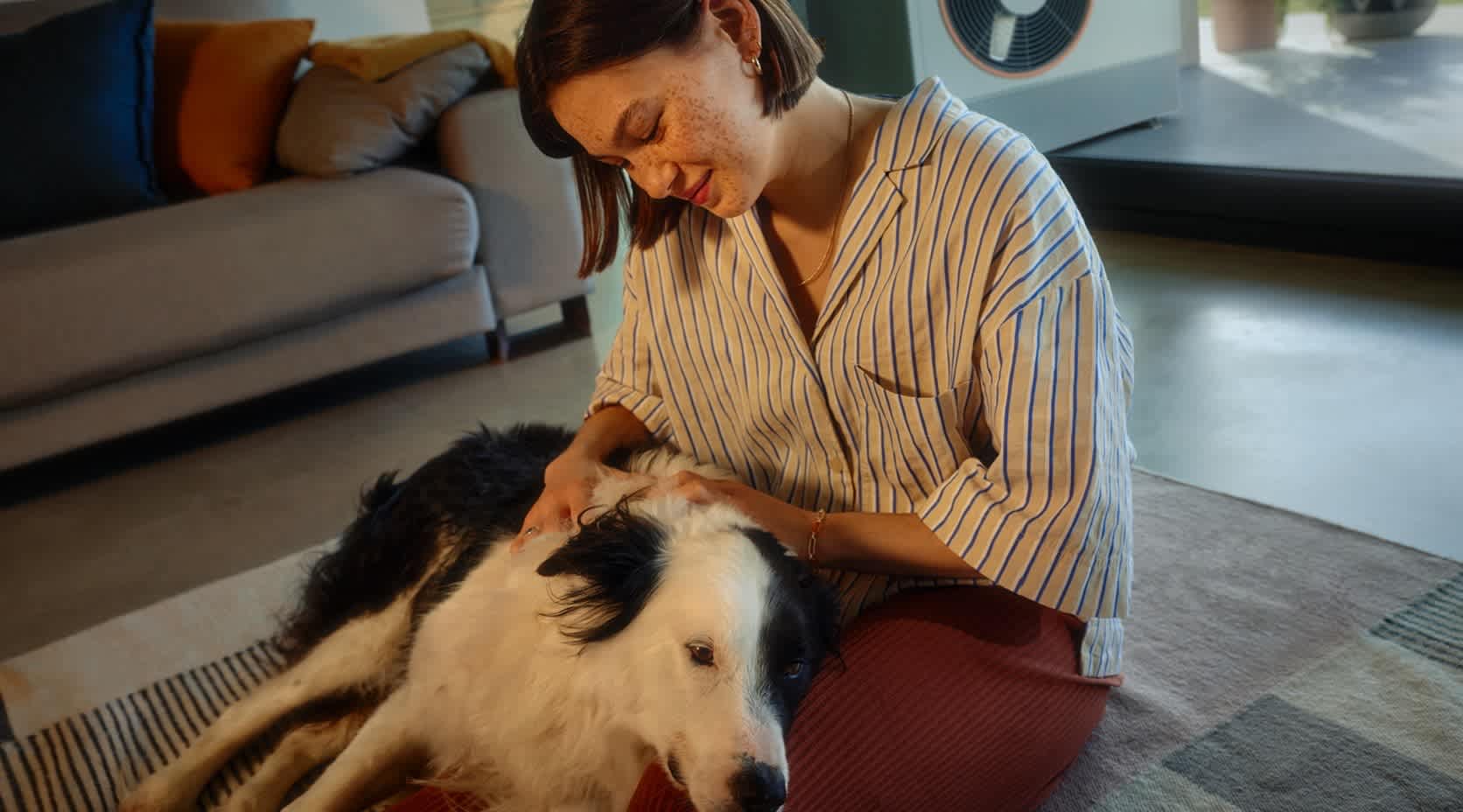 Junge Frau liegt mit ihrem Hund auf einem Teppich im Wohnzimmer ihres Hauses. Im Hintergrund auf der Terrasse ist eine Wärmepumpe  zu sehen.