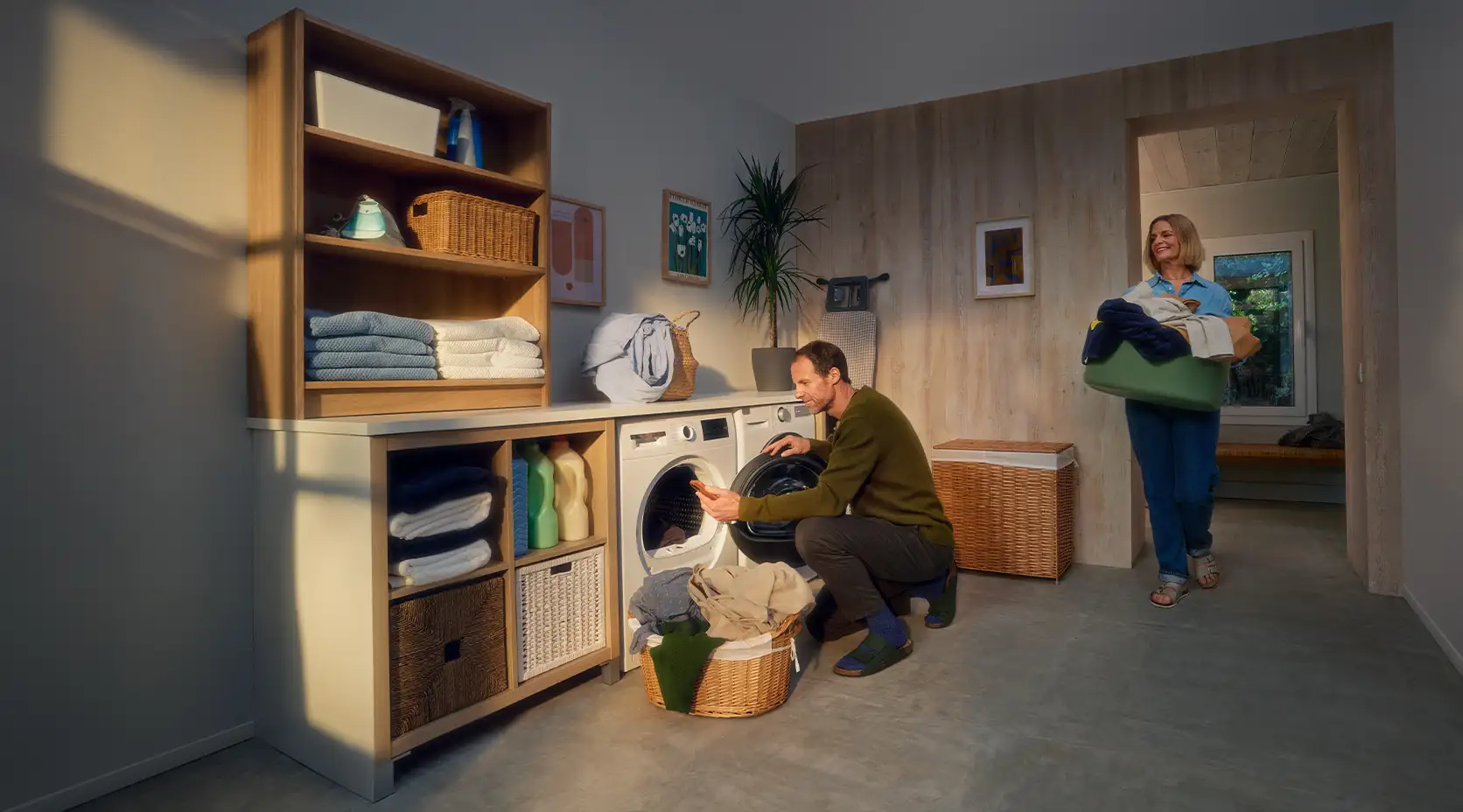 Ein Mann befüllt eine Waschmaschine und seine Frau bringt mehr Wäsche im Wäschekorb.