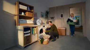 Ein Mann befüllt eine Waschmaschine und seine Frau bringt mehr Wäsche im Wäschekorb.