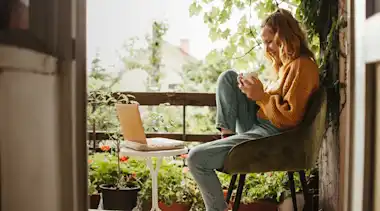 Junge Frau sitzt mit Tasse vor Laptop auf Balkon