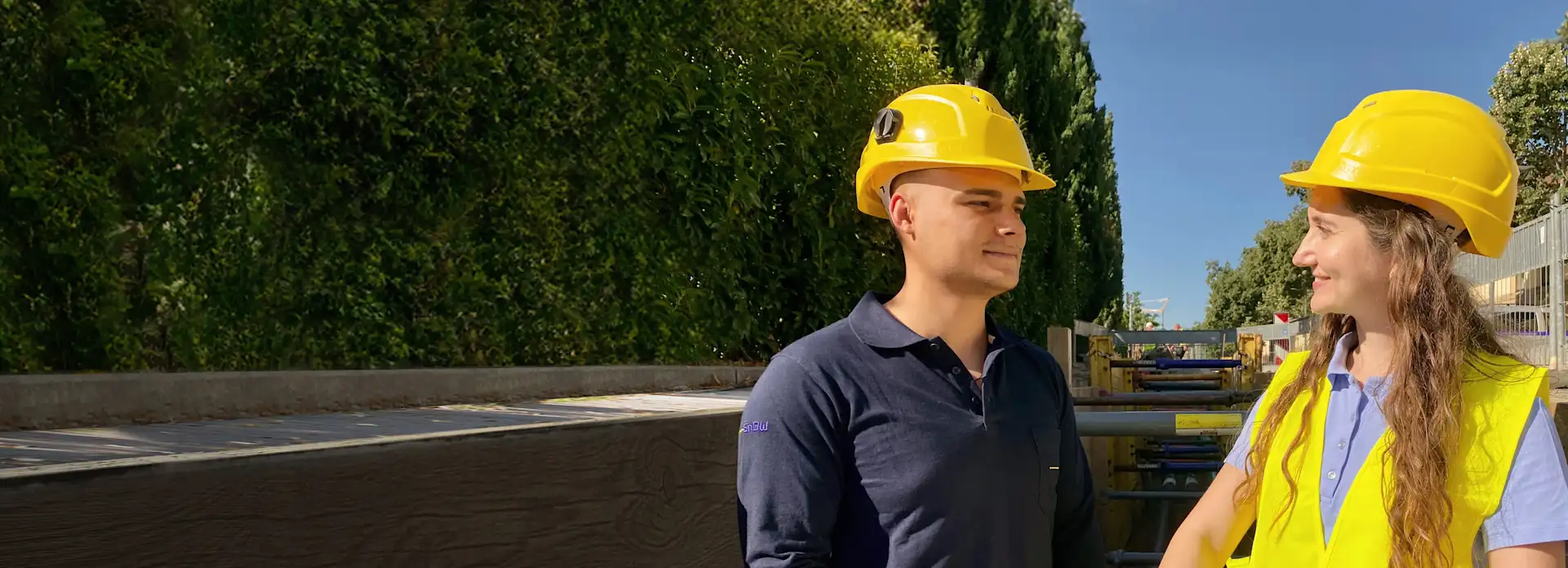 Mann und Frau mit Bauhelmen vor Hecke blauer Himmel