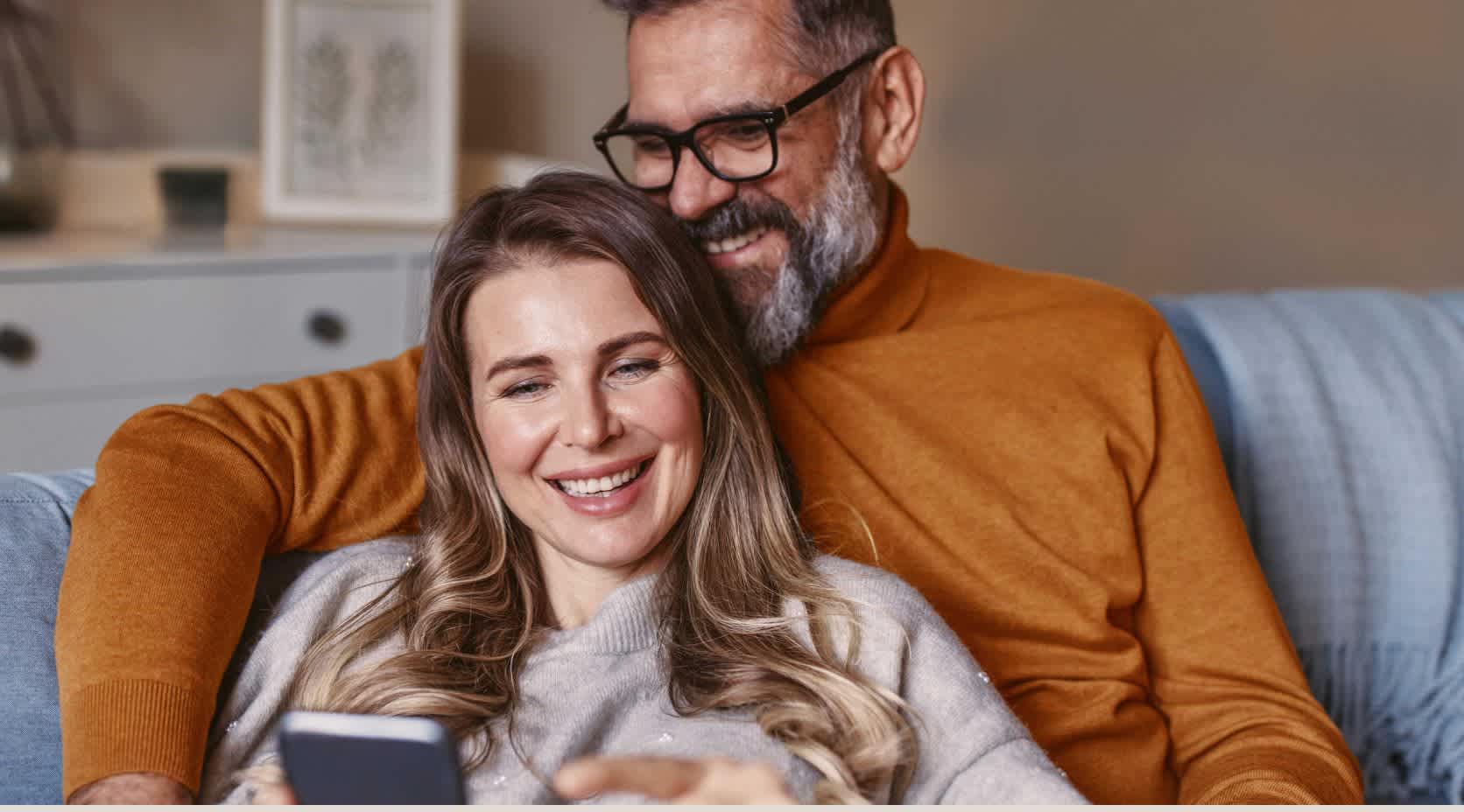 Pärchen sitzt gemütlich auf der Couch und schaut auf ein Smartphone.