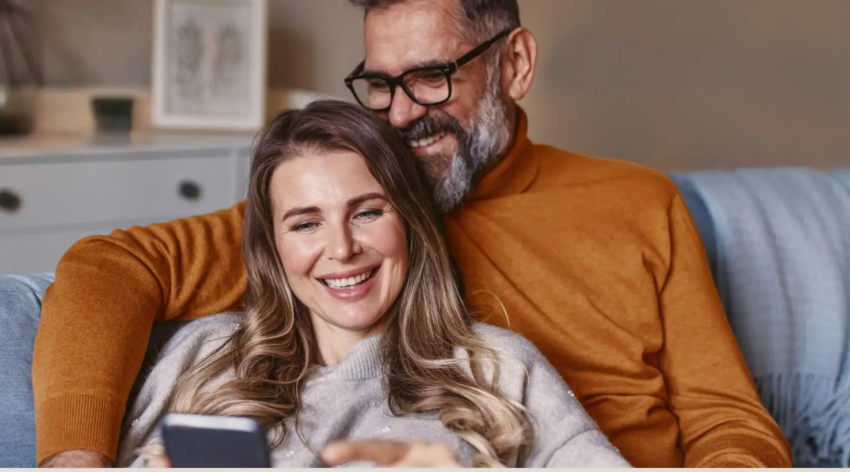 Pärchen sitzt gemütlich auf der Couch und schaut auf ein Smartphone.