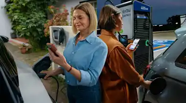 Zwei Frauen laden ihr Auto zuhause und unterwegs und prüfen den Ladevorgang in der EnBW mobility+ App auf ihrem Smartphone