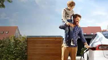 Vater hat Sohn mit Brille auf dem Arm beim Elektroauto Laden Wohnsiedlung