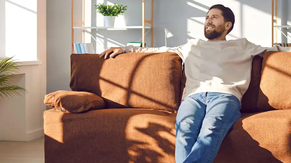 Mann sitzt lächelnd entspannt aus dem Sofa neben Palme im Wohnzimmer