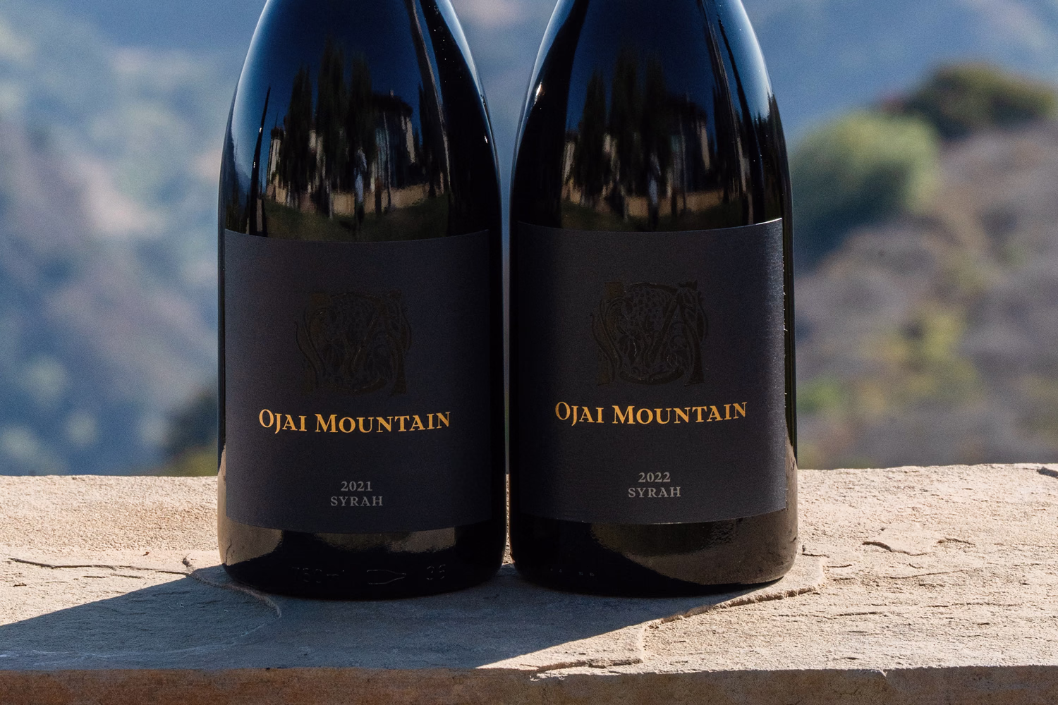 Ojai Mountain 2021 and 2022 Syrah bottles on a stone ledge above the vineyard hills Ojai Mountain 2021 and 2022 Syrah bottles on a stone ledge above the vineyard hills
