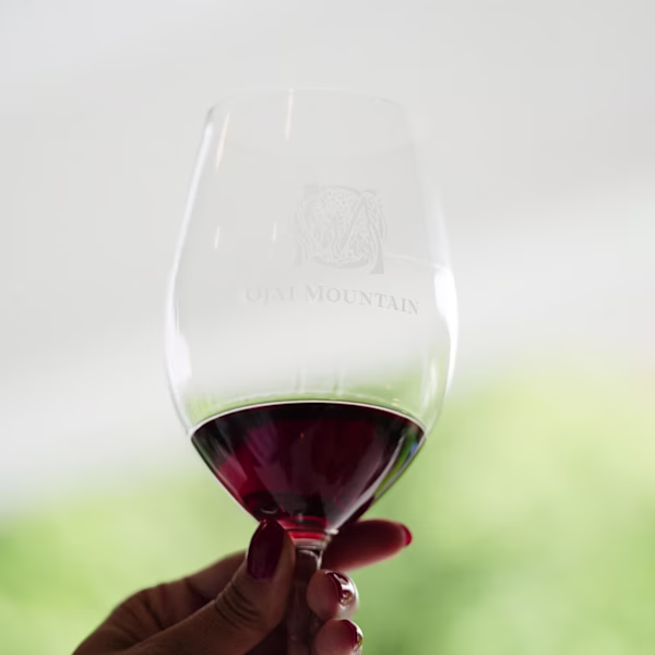 Swirling Ojai Mountain Red Wine in the Vineyard Hand holding a glass of red wine with Ojai Mountain logo against green vineyard bokeh
