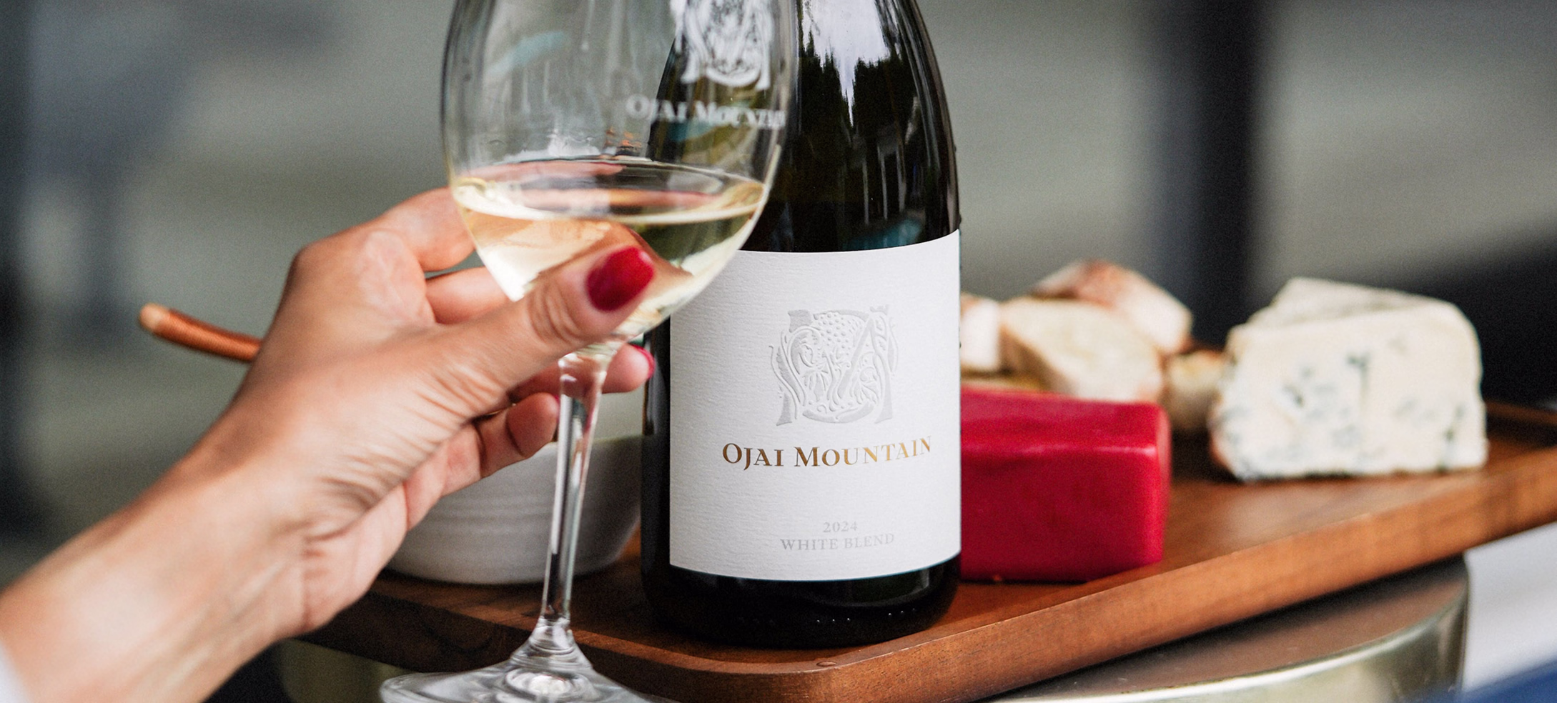 Hand holding white wine beside an Ojai Mountain bottle and cheese board Hand holding white wine beside an Ojai Mountain bottle and cheese board
