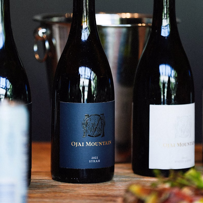 Ojai Mountain 2021 Syrah bottles beside a silver ice bucket on wood