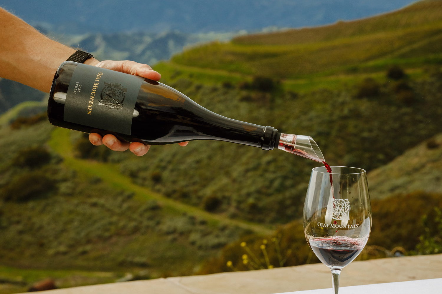 Ojai Mountain 2022 Syrah poured into a branded glass above the vineyard hills Ojai Mountain 2022 Syrah poured into a branded glass above the vineyard hills