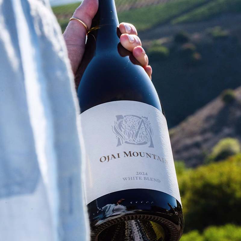 Hand holding Ojai Mountain 2024 White Blend wrapped in cloth above the hills