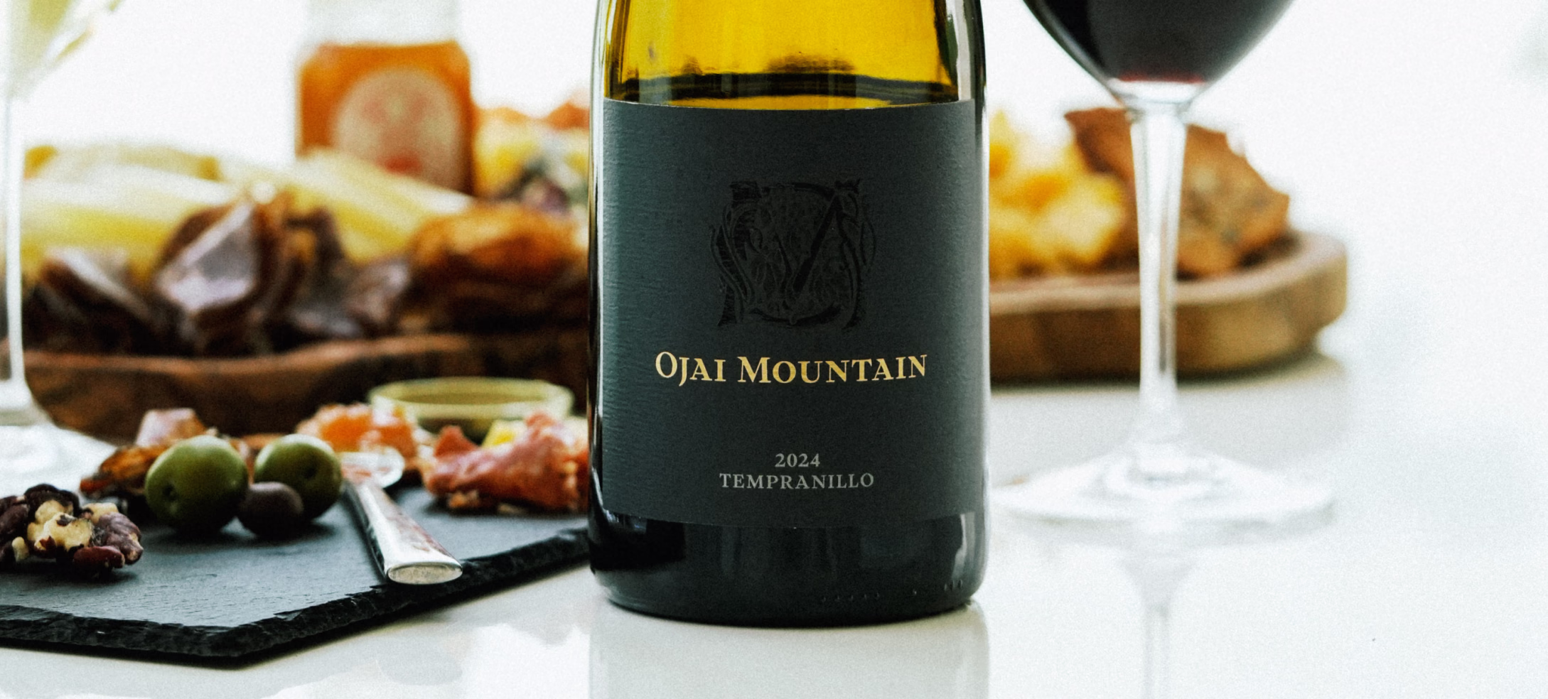 A bottle of Ojai Mountain 2024 Tempranillo beside olives, cheese, a charcuterie board, and a glass of red wine.