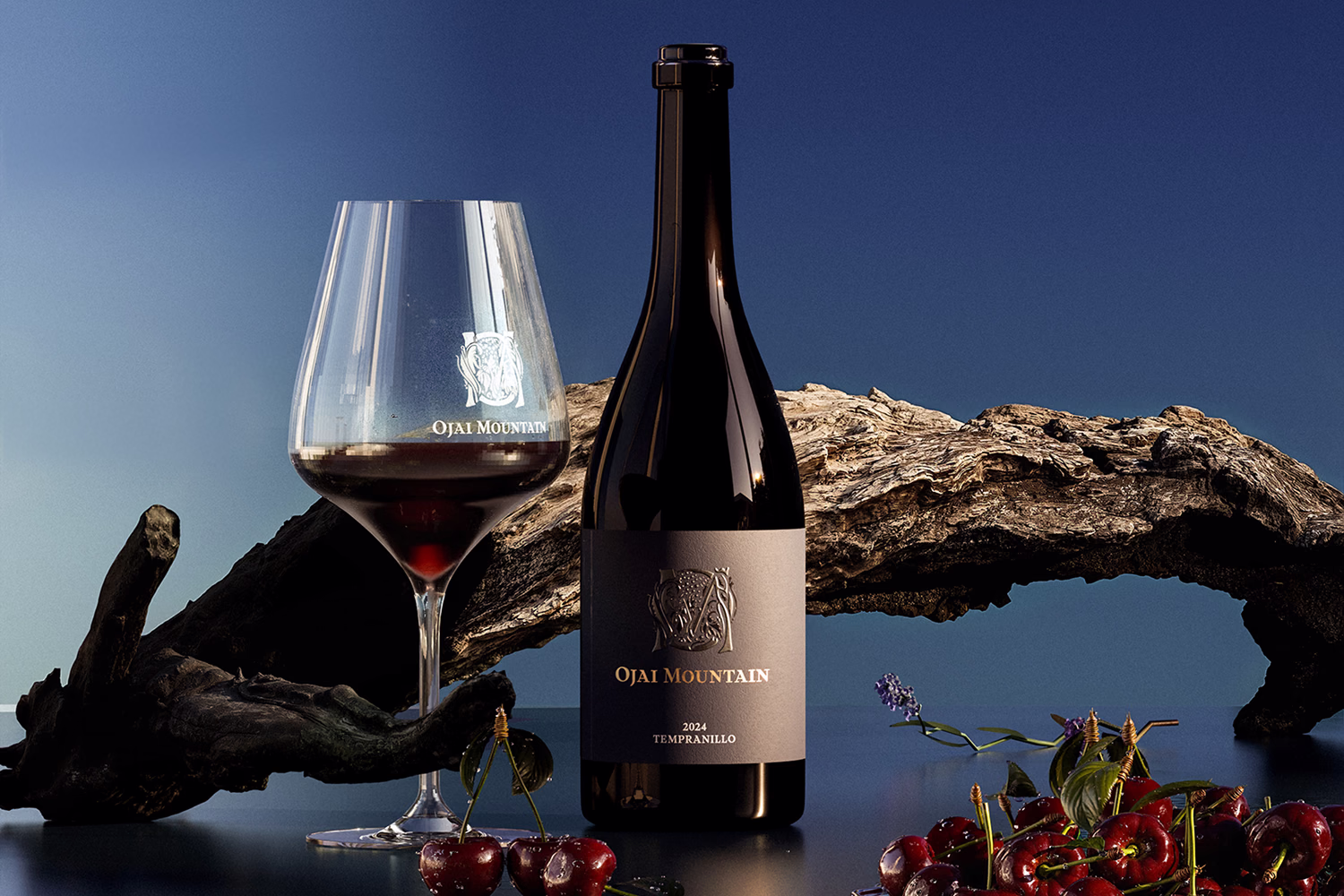 Ojai Mountain 2024 Tempranillo bottle and glass beside driftwood with cherries and a blue backdrop