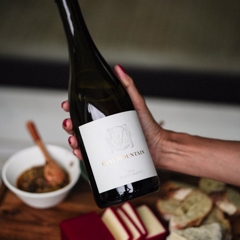 Hand holding Ojai Mountain 2024 White Blend above a table of food