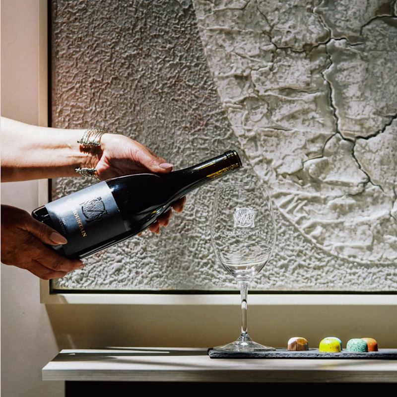 Hand pouring Ojai Mountain Tempranillo into a glass beside a textured plaster wall