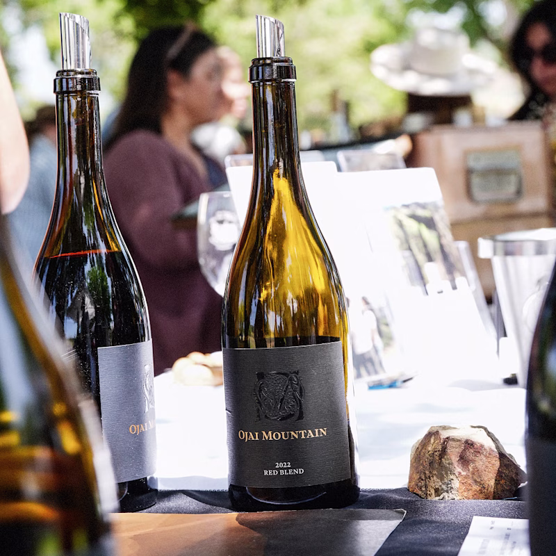 Ojai Mountain bottles on a table with guests gathered outdoors behind Ojai Mountain bottles on a table with guests gathered outdoors behind