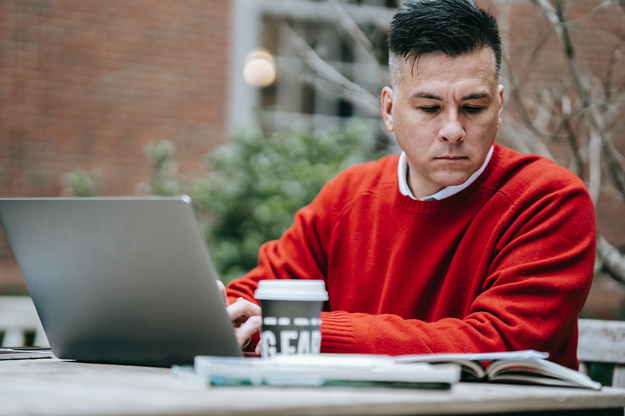 Person in red jumper looks perplexed at piece of paper whilst using a laptop on an outdoor cafe table