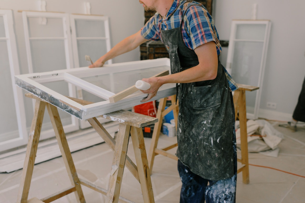 person in apron and check shirt painting window frames resting on wooden trestles in a garage