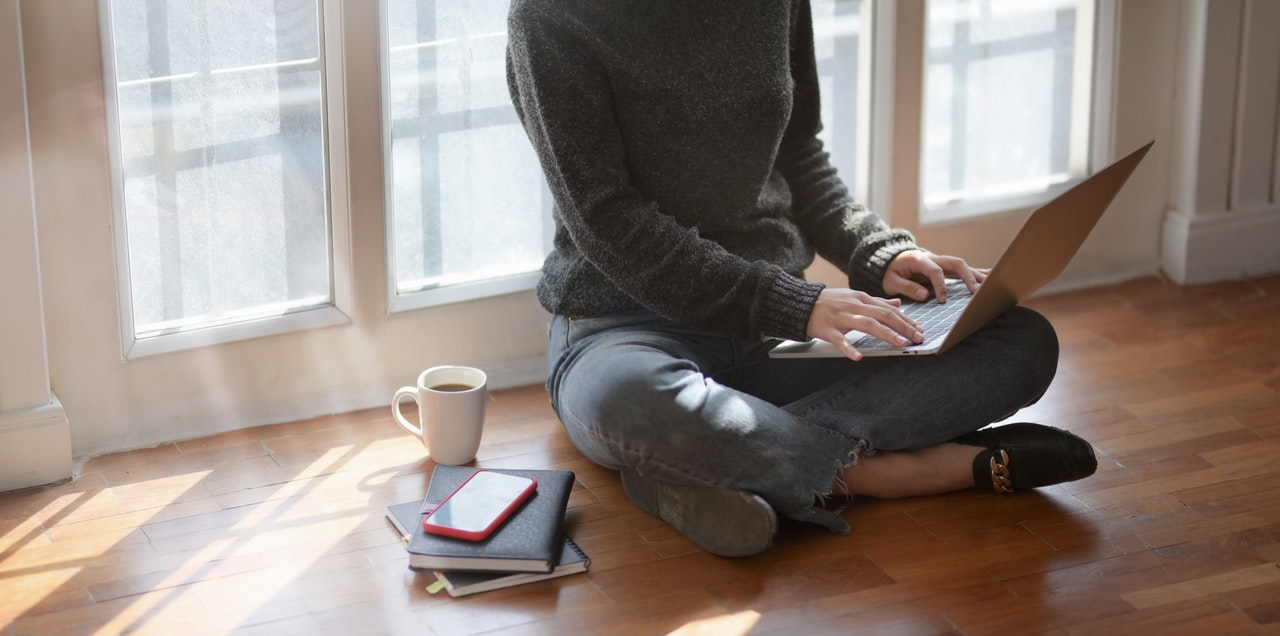 person sat on wooden floor using laptop, pile of notepads on floor next to them